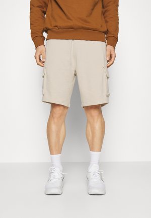 Mann trägt beige Cargo-Shorts, weiße Socken, weiße Sneakers und ein braunes Langarmshirt, steht vor einem einfarbigen Hintergrund.