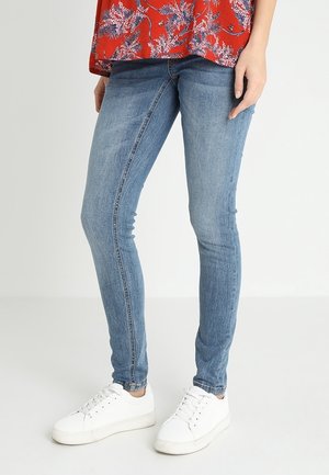 Person iført smalle blå jeans, hvide sneakers og en rød blomstret top, stående mod en ensartet lys baggrund.