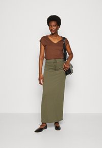 Brown V-neck t-shirt paired with a high-waisted olive green maxi skirt. The model wears black flat shoes and holds a textured bag.