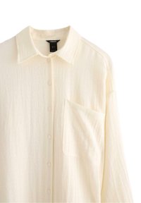 Camicia button-up di colore crema chiaro realizzata in un tessuto testurizzato, con colletto e tasca sul petto. Presenta sette bottoni lungo la parte anteriore.