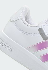 Bílá syntetická teniska s texturovaným povrchem, růžovými iridiscentními pruhy, polstrovaným límcem a reliéfním značením na podrážce.