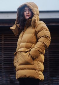 Chaqueta tipo puffer en color mostaza, con capucha, cintura ajustada y bolsillos laterales, con textura acolchada y acabado brillante.