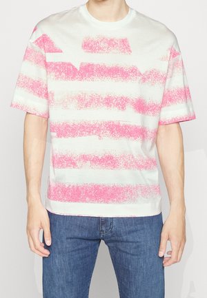 Osoba ubrana w biały t-shirt z różowymi poziomymi paskami w stylu pociągnięć pędzla i niebieskie dżinsy, stojąca na jednolitym tle.