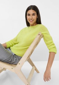 Limegrüner Strickpullover mit langen Ärmeln, getragen über grauen Nadelstreifenhosen, sitzt auf einem hellen Holzklappstuhl. Minimalschmuck.