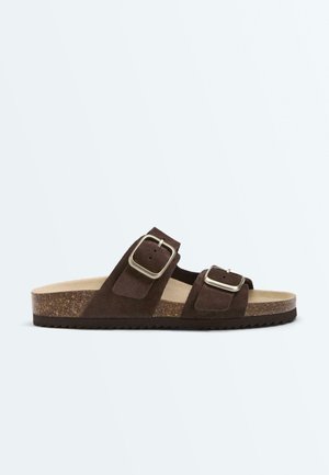 Sandalo tipo slide in camoscio marrone con due fibbie regolabili color argento, sottopiede in sughero e suola nera scanalata, mostrato di lato su sfondo bianco.