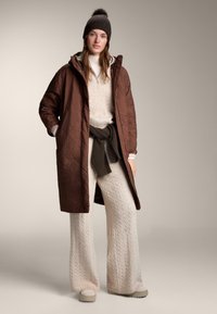 Hnedý prešívaný dlhý kabát cez krémový sveter s vrkočovým vzorom a široké nohavice, doplnené tmavou pletenou čiapkou a svetlými topánkami.