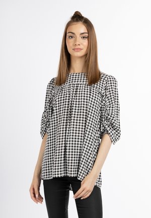 Jonge vrouw staat, draagt een zwart-wit geblokte blouse met geplooide mouwen en zwarte broek tegen een effen witte achtergrond.