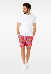 Red patterned swim shorts with various cartoon graphics, white drawstring waist, worn with a plain white t-shirt and white sneakers.