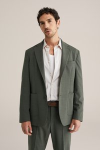 Bărbat în sacou și pantaloni gri, purtând o cămașă albă cu guler deschis și o curea maro, stând pe un fundal simplu.