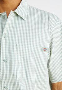 Světle modrá a bílá kostkovaná košile s krátkým rukávem, s límečkem na knoflíky, přední kapsou a nášivkou s logem. Měkký bavlněný materiál.