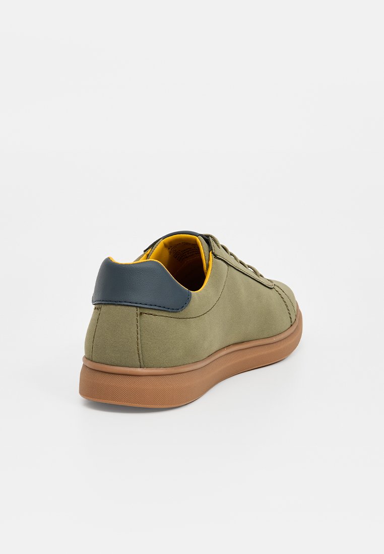 Olivgrüne Sneaker mit brauner Sohle, marineblauem Fersenbereich und gelbem Innenfutter, von hinten auf einem weißen Hintergrund betrachtet.