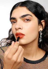 Modèle appliquant un rouge à lèvres orange profond, portant un top en maille noire avec des détails irisés et de grandes boucles d'oreilles argentées.