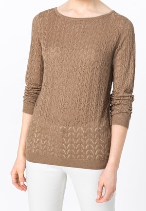Femme portant un pull en maille texturée marron clair à manches longues et un pantalon blanc, montré des épaules jusqu'aux hanches.