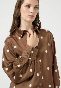 Chemise marron à pois en tissu lisse, avec un col à revers et des poignets à boutons. Coupe ample avec manches plissées.