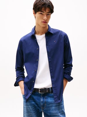 Conjunto informal que incluye una camisa ajustada de botón en azul marino, una camiseta blanca y jeans azules. Los detalles notables incluyen un logo en la camisa y un cinturón negro.