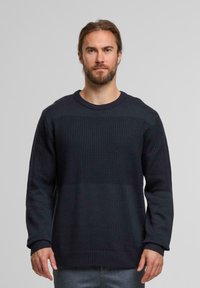 Uomo con barba e capelli raccolti, che indossa un maglione a maglia scura e trapuntata e pantaloni grigi, in piedi contro uno sfondo chiaro e uniforme.