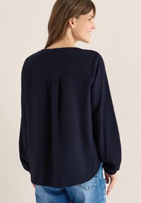 Blouse marine à coupe ample, manches longues et col rond. Présente un pli au dos et des poignets élastiques. Associée à un jean bleu clair.