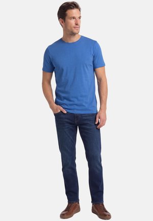 Mies seisoo valkoista taustaa vasten, toisessa kädessä taskussa, yllään sininen lyhythihainen t-paita, tummat farkut ja ruskeat nahkakengät.