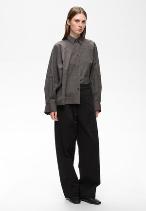 Femme aux cheveux lisses porte une chemise à carreaux oversize, un pantalon large noir et des chaussures noires, debout devant un fond blanc uni.