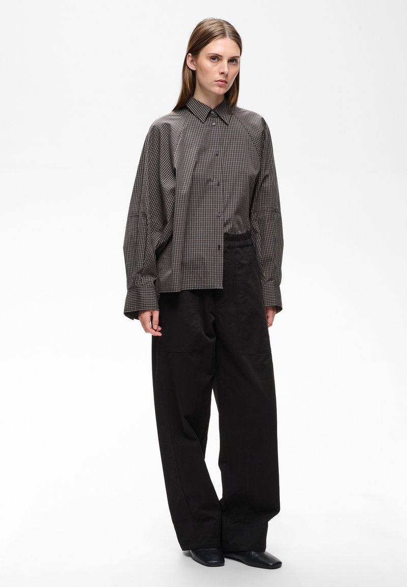 Femme aux cheveux lisses porte une chemise à carreaux oversize, un pantalon large noir et des chaussures noires, debout devant un fond blanc uni.