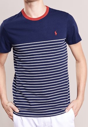Homme portant un t-shirt à manches courtes bleu marine et blanc à rayures avec un col rouge et un logo rouge sur la poitrine, les mains dans les poches d'un pantalon blanc.