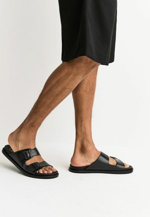 Male legs in black shorts wearing black slide sandals with double buckle straps on left foot and single wide strap on right foot.
