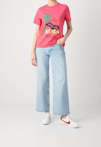 Camiseta de manga corta rosa con un gráfico de "BEACH", combinada con jeans de pierna ancha en azul claro y zapatillas blancas con acentos rojos.