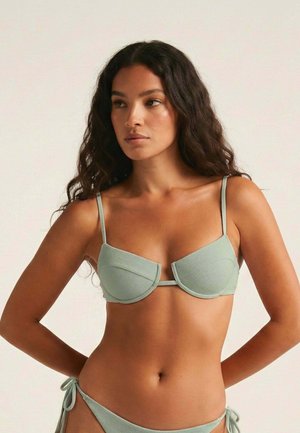 Mujer con cabello largo y rizado que lleva un bikini texturizado de color verde claro, mirando hacia un lado contra un fondo claro y liso.