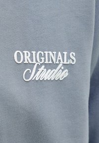 Gaiši zils austs audums ar uzpūstu baltu izšūtu tekstu "ORIGINALS Studio".