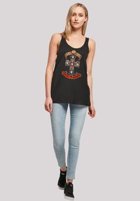Schwarzes Tanktop mit einem farbenfrohen Grafikdesign, das ein Totenkopf-Motiv und Text zeigt. Kombiniert mit hellblauen Skinny-Jeans und schwarzen Sneakers.