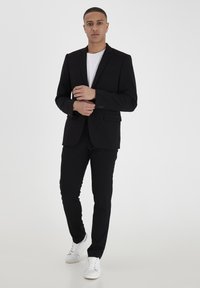 Hombre de pie vestido con un traje negro, camiseta blanca y zapatillas blancas sobre un fondo blanco liso.