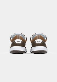 Bruna mocka- och kanvassneakers med en vit gummisula. Bakåtvyn visar logotypen på hälen och kontraster mellan ljus och mörkbrun.