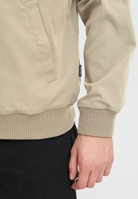 Veste bomber beige avec des poignets et un ourlet côtelés, dotée d'une poche latérale et d'un tissu texturé. Jean noir partiellement visible à l'arrière-plan.