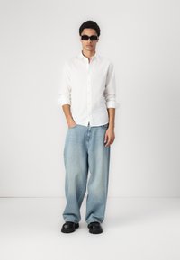 Hvid linnedskjorte, lyseblå oversized jeans og sorte sko med tykk sål. Model iført mørke solbriller, stående med hænderne i lommerne.