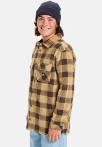 Braun und beige karierter Flanellhemd mit zwei Brusttaschen und einem schwarzen.logo-patch. Gestylt mit einer dunklen Mütze. Lässiger, entspannter Schnitt.