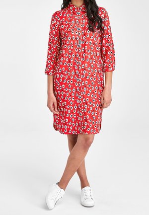 Femme portant une robe-chemise rouge à motifs longueur genou avec des manches trois-quarts et des baskets blanches, debout les jambes croisées.