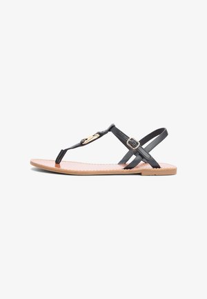Flat sandal med tan såle og svarte lærremmer, med en tåsløyfe, justerbar ankelspenne, og en liten gullfarget dekorativ plate på den fremre remmen.