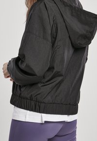 Zwarte, waterdichte jas met een capuchon, elastische manchetten en een gerimpelde tailleband. Gladde textuur en lichtgewicht ontwerp. Achteraanzicht weergegeven.