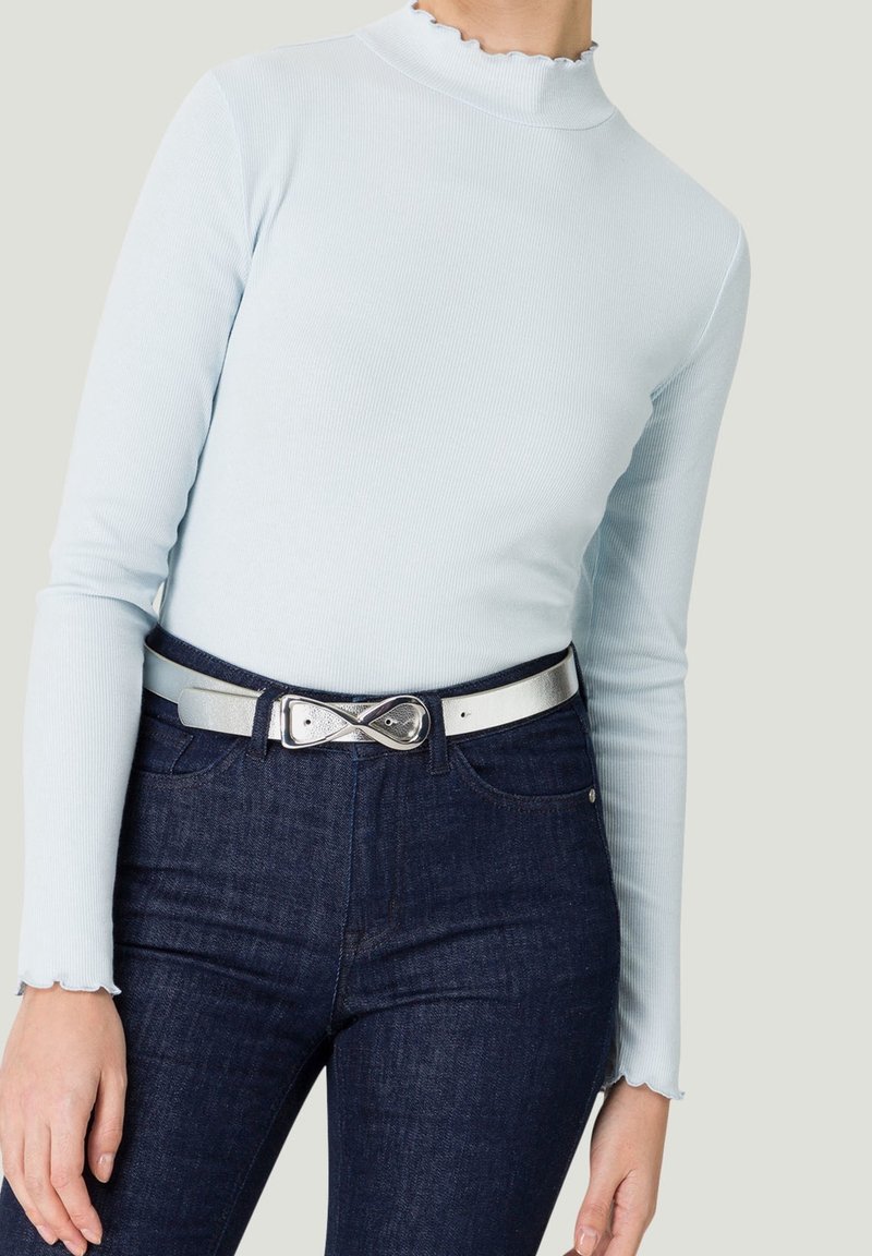 Femme portant un haut à manches longues côtelé bleu clair avec des bords ondulés aux poignets et à l'encolure, un jean bleu foncé et une ceinture argentée avec une boucle en forme d'infini.