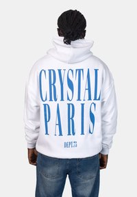 Felpa bianca con grande testo blu sul retro: "CRYSTAL PARIS" e "DEPT.75." Il tessuto sembra morbido; il design è oversize con cappuccio.