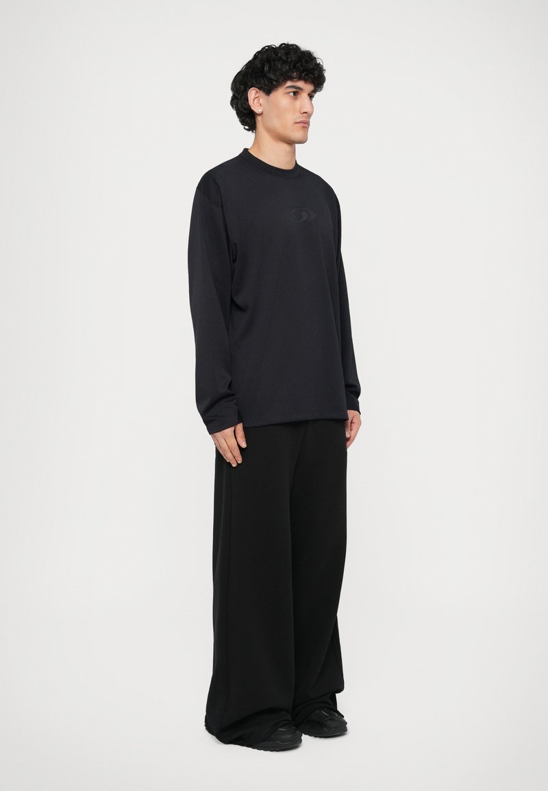 Maglietta nera a maniche lunghe con un motivo testurizzato, abbinata a pantaloni larghi neri. L'outfit presenta dettagli minimi e una silhouette rilassata.