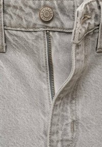 Gros plan d’un jean en denim gris clair avec une fermeture éclair partiellement ouverte et un bouton métallique marqué de texte et de chiffres.