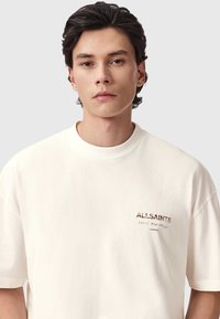 Μπλούζα σε κρεμ απόχρωση, με μεγάλα μεγέθη και στρογγυλή λαιμόκοψη, κατασκευασμένη από βαμβάκι, με λογότυπο και καφέ κείμενο στην αριστερή πλευρά του στήθους. Λεία υφή.