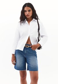 Camisa blanca de botones con cuello, combinada con pantalones cortos de mezclilla azules con bordes deshilachados. Los accesorios incluyen un cinturón de patrón y un bolso pequeño.