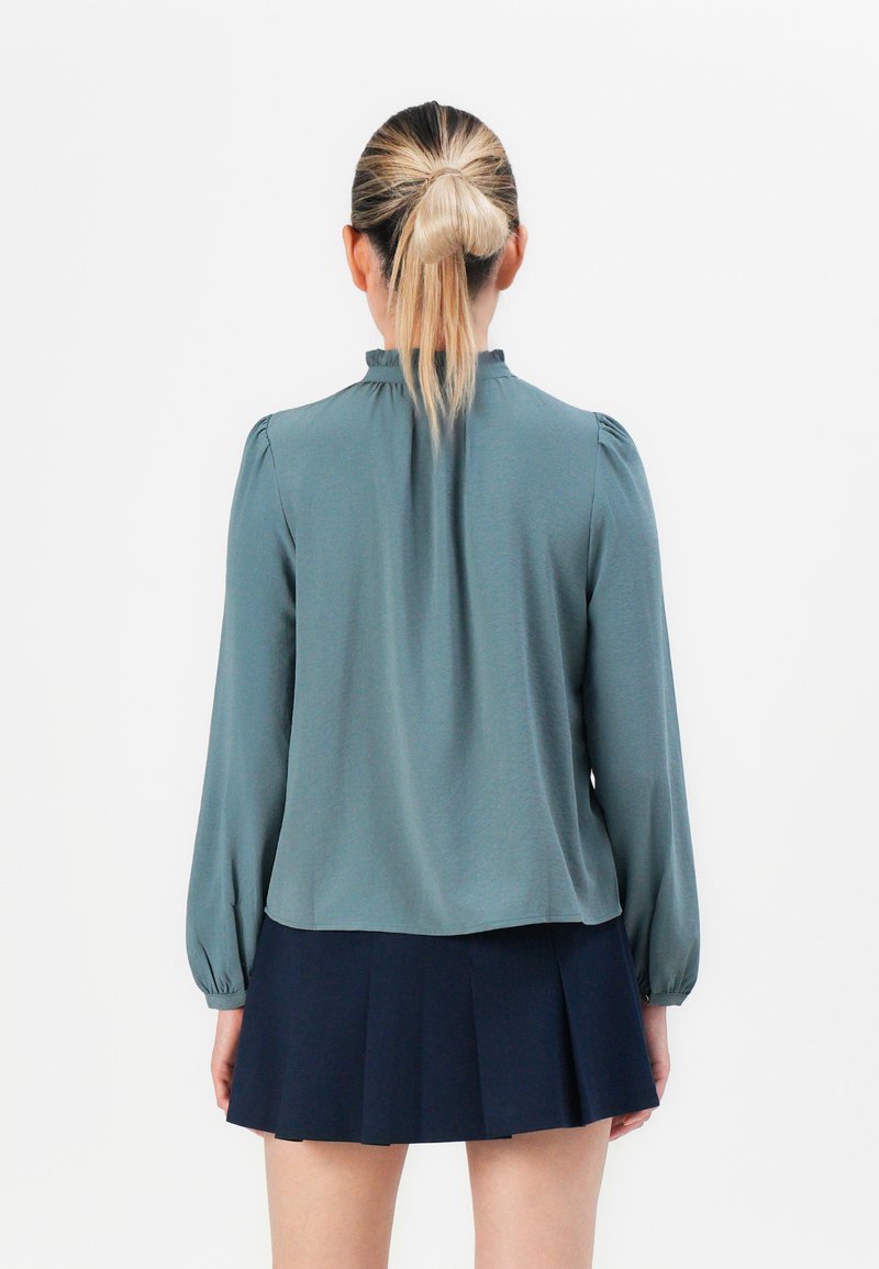 Vrouw met een blonde paardenstaart, gekleed in een blauwe blouse met lange mouwen en een donkere geplooide rok, staand voor een effen witte achtergrond.