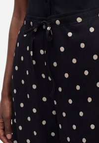 Pantalon noir à pois blancs, en tissu texturé avec taille à cordon noué, porté par une personne à la peau foncée.