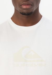 Närbild av en mans undre ansikte och hals som bär en vit T-shirt med en svag Quiksilver-logga på bröstet.