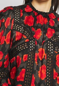 Schwarze Bluse aus strukturiertem Stoff mit roten Blumenmustern, Spitzendetails und einem hohen Kragen mit gerafften Abschlüssen.