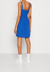 Vestido azul sin mangas con cuerpo fruncido, falda acampanada y tirantes ajustables. Llevado con zapatillas blancas y calcetines, mostrado desde atrás.