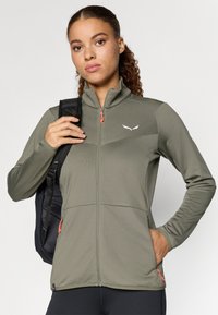 Olivgrüne Zip-Jacke aus strukturiertem Stoff, mit orangefarbenen Akzenten, hohem Kragen und zwei Fronttaschen mit Reißverschlüssen.
